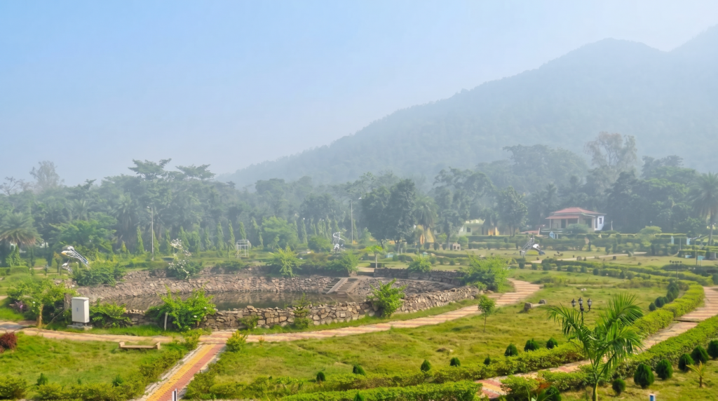 A scenic view of rustic eco-resorts and red-tiled cottages nestled at the base of Susunia Hill, surrounded by a lush forest of Sal and Palash trees under a clear sky.